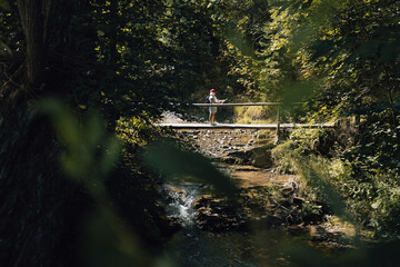 Vertical photo of a person standing quietly on a small wooden bridge in a dense, sun-dappled...