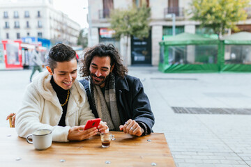 Gay couple smiling and using smartphone at outdoor cafe