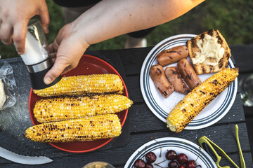 Delicious barbecue spread with grilled corn, sausages, cherries, ready for a summer gathering. A person seasons grilled corn on the cob on a black outdoor table, ideal for a summer barbecue party.