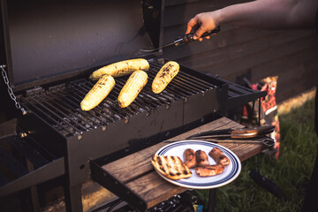 Grilled corn on the cob and sausages are ready for a summer barbecue party. A person grilling corn...