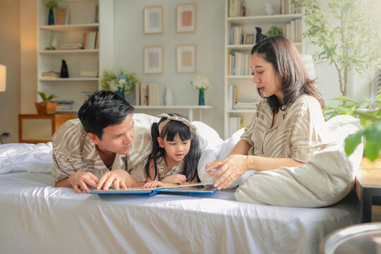 Happy Asian family lying together in bed, with mother and daughter cuddling under a warm blanket while father reads a bedtime story from a book. Parents and child bonding through storytelling cozy