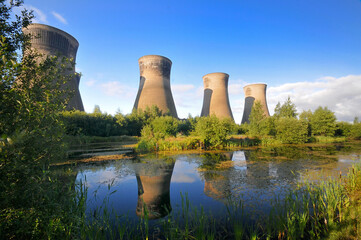 Disused cooling towers reflected in a tranquil pond, surrounded by lush greenery under a clear sky