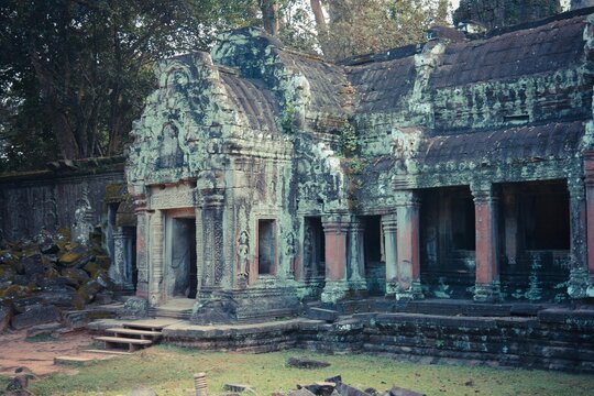 Khmer temple structure with intricate carvings, darkened stone, and collapsed sections amid green moss and fallen rocks.