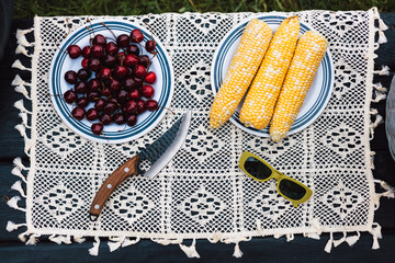 A summer picnic scene with fresh cherries and corn on the cob plate. A rustic picnic scene featuring cherries, corns, a knife, and stylish green sunglasses on a decorative white vintage tablecloth.