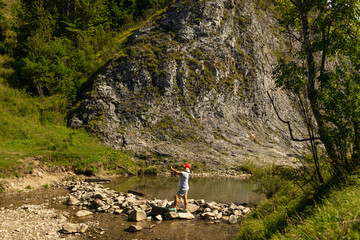 Active senior woman in casual hiking clothes stepping over stones near a calm mountain pond...
