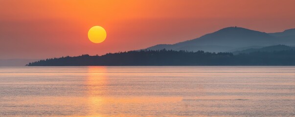 Obraz premium A vibrant sunset over calm waters with silhouetted hills in the background, casting an orange glow across the serene landscape.