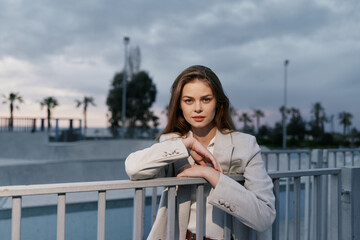 Woman in light suit leaning on metal railing outdoors with cloudy sky and palm trees in background,...