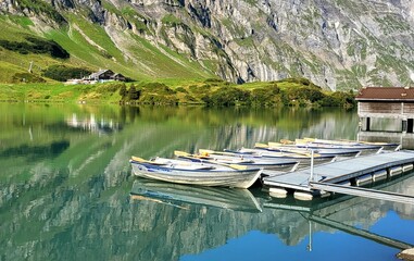 Naklejka premium Boote an einem Steg beim Bergsee Trübsee mit türkisblauem Wasser und atemberaubender Spiegelung sowie stahlblauem Himmel in der leuchtend klaren Morgensonne, Trübsee, Engelberg, Schweiz