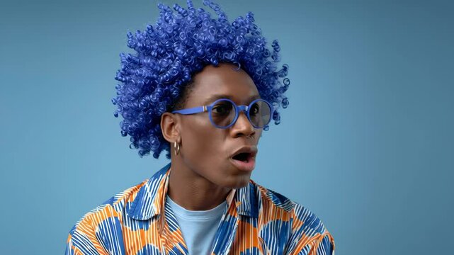 Man with blue hair and glasses is wearing a colorful shirt. He is looking at the camera with a serious expression