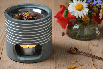 Incense burner with tealight and summer flowers in a vase on wooden table. Dried herbs, resin and...