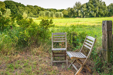 Two weathered folding chairs at the edge of a meadow. Both chairs are empty. Around the grassland...