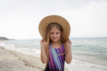 Portrait of cute little girl in swimwear and straw hat standing on the sandy beach with blue sea...