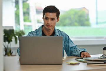 Young Asian Graphic Designer Man Concentrating on Laptop in Modern Office