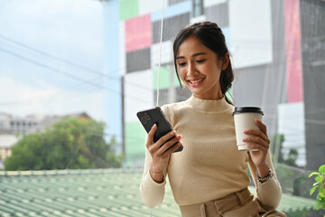 Confident businesswoman in casual office attire holding a cup and phone, taking a break in a bright...