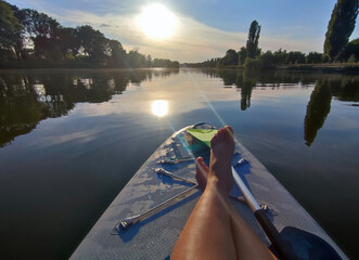 Bare legs of woman on SUP board on surface of river or lake with sun reflecting and with trees on...