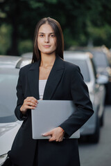 Woman in business suit stands with laptop near her car