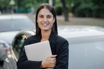 Woman in business suit holding laptop leaning on her car