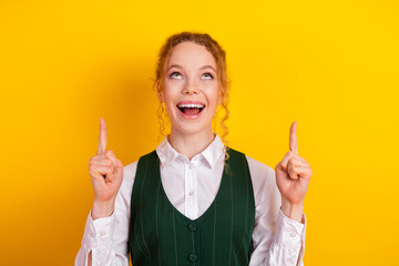 Young woman pointing upwards on bright yellow background wearing a green vest and smiling...