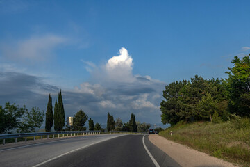 An asphalt road in Crimea, curving among green hills and cypresses against a dramatic sky with...