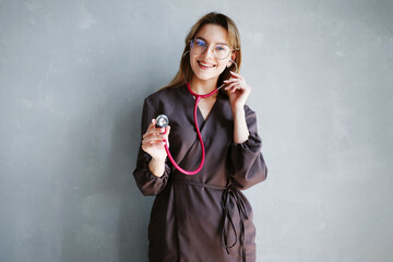 Portrait of female doctor in dark medical uniform smiling and using stethoscope while standing against gray wall.