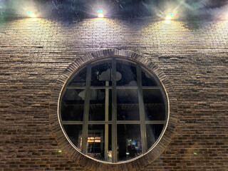 Snowfall creates a serene atmosphere outside a round window of a brick building at night