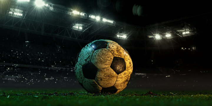 Worn soccer ball on grass in the middle of a 3D-rendered stadium at night with floodlights in the background. Concept of positioning, anticipation, and spatial awareness