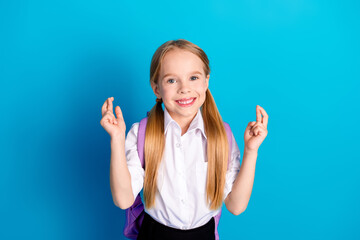 Smiling young schoolgirl in white uniform on blue background making a crossed fingers gesture while...