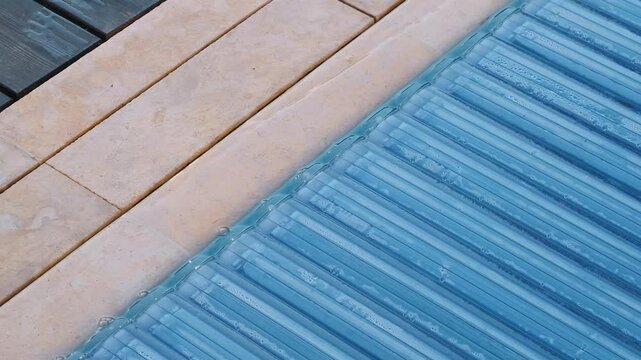 Closed pool surface with an automatic shutter and water droplets, adjacent to stone-tiled coping. A detailed close-up with a geometric pattern
