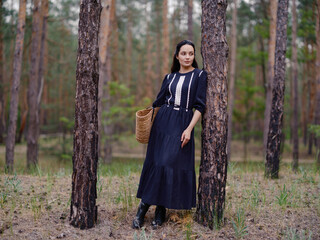 Young Woman in dark purple Dress with Straw Bag Standing among pine trees in forest.