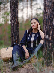Young woman in dark purple dress sitting among pine tree trunks in forest next her straw bag.