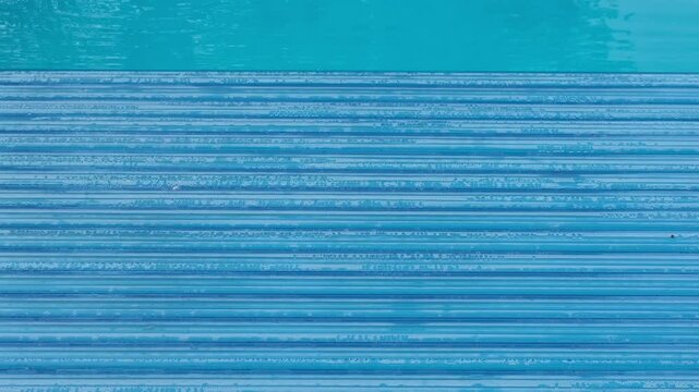 Swimming pool covered with an automatic shutter, water droplets on the surface. Top-down view, the panel texture forms a rhythmic pattern in cool blue tones
