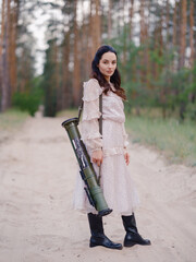 Young woman in light dress standing on sandy forest path and holding grenade launcher.