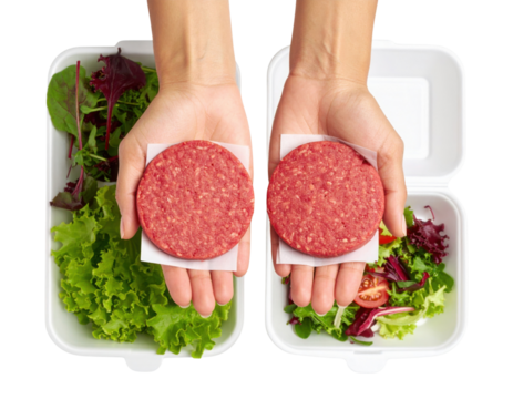 Two raw beef burger patties held in hands above containers filled with fresh mixed salad greens and sliced tomatoes.