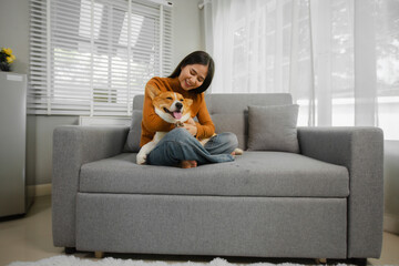Young Asian woman sitting with her dog on the sofa at home with headphones and playing on laptop on holiday.