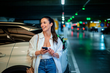 Woman leaning against her car in a parking garage and using a smart phone