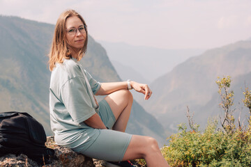Young woman in sportswear sits on a rock ledge, peacefully observing scenic mountain landscape during a summer hiking adventure.