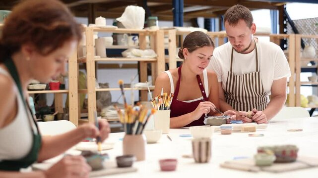 Experienced potter helps students learn how to make patterns on ceramic plates using paint and brush in a pottery workshop. High quality 4k footage