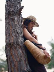 Young woman in brown dress and straw hat holding straw bag, standing against background of pine...