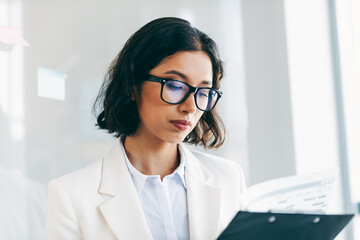 Professional businesswoman in formal attire reading documents in an office environment with natural...