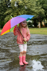 Сheerful little girl stands in a puddle with a colorful umbrella during a summer rain. Toddler...