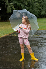 Little girl with a transparent umbrella and rubber boots walks in rainy weather in summer. Happy...