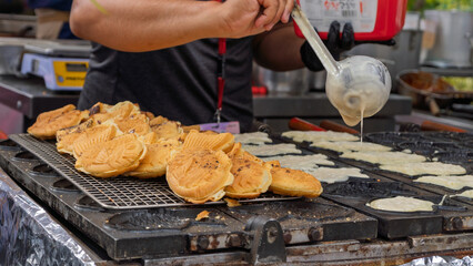 Taiyaki being prepared at street food stall with traditional griddle. Outdoor culinary scene with cooks in action.