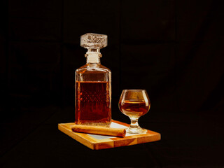 Elegant whiskey decanter and glass served on a wooden tray with a rolled cigar, set against a dark...