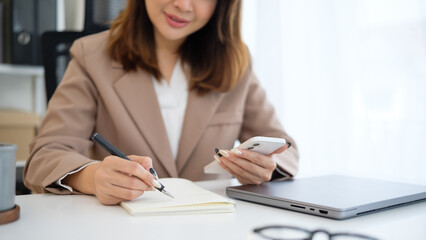 Smiling businesswoman writing notes while checking her smartphone at the office.