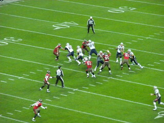 American football game in action between two teams on a grassy stadium field, with players in red and white uniforms and referees observing play © Eniko
