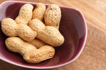 Unpeeled roasted peanuts in a red ceramic bowl on wooden brown surface. Ready to eat nut snack. Macro shot