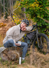 Cute young female cyclist enjoying peaceful break in autumn forest. Sitting on log, holding cup and...