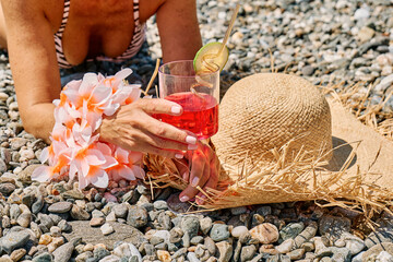 Anonymous woman in bikini ad straw hat holding summer refreshing red soda drink on pebble beach.