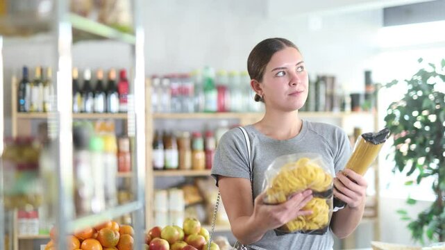 Youth girl choose and buy pasta in store. Market visitor consider and make choice of assortment, buy noodles. Client hesitates before buying, choose garnish product, reads package labels 