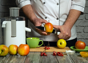 A person in a chef's uniform slices an apple to create fresh juice. Juicer, fruits, green cup, and...